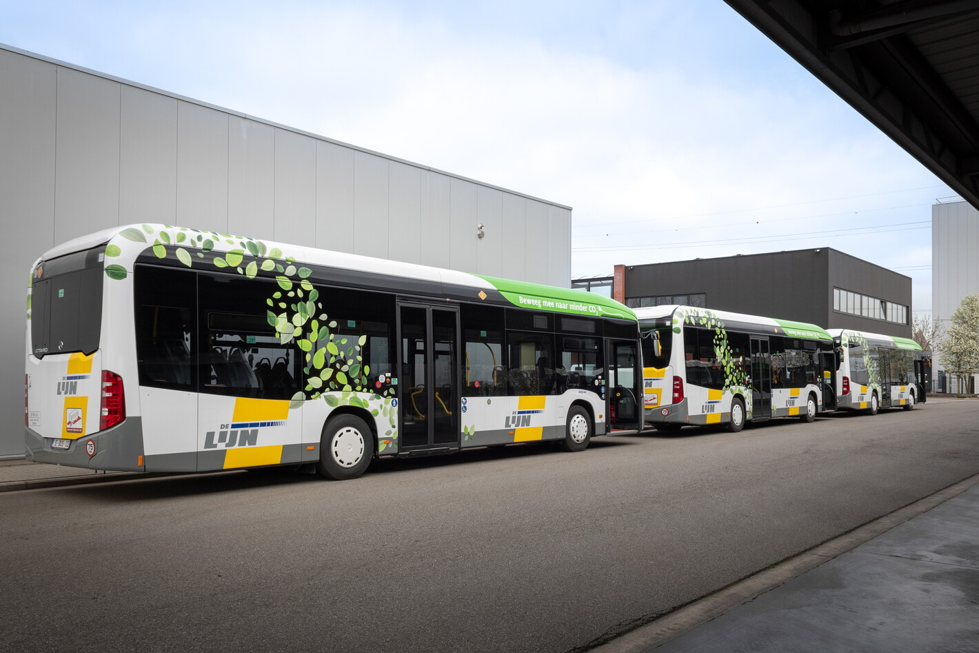 Drie bussen met geel-groen patroon geparkeerd bij een depot op een zonnige dag