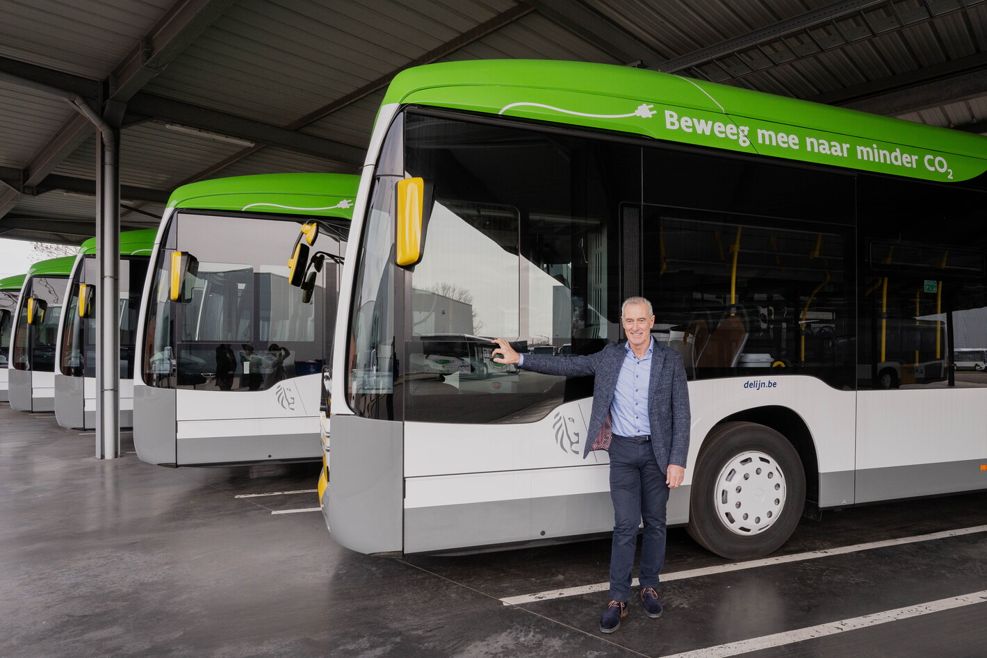 Man in pak staat voor een rij witte bussen met groene accenten in een depot.