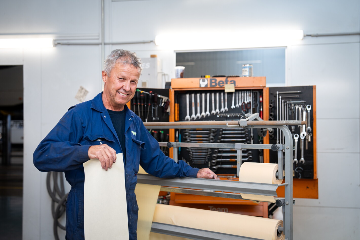 Oudere man in werkplaats bij houtbewerkingsmachine, houdt lichtgekleurde plank vast.