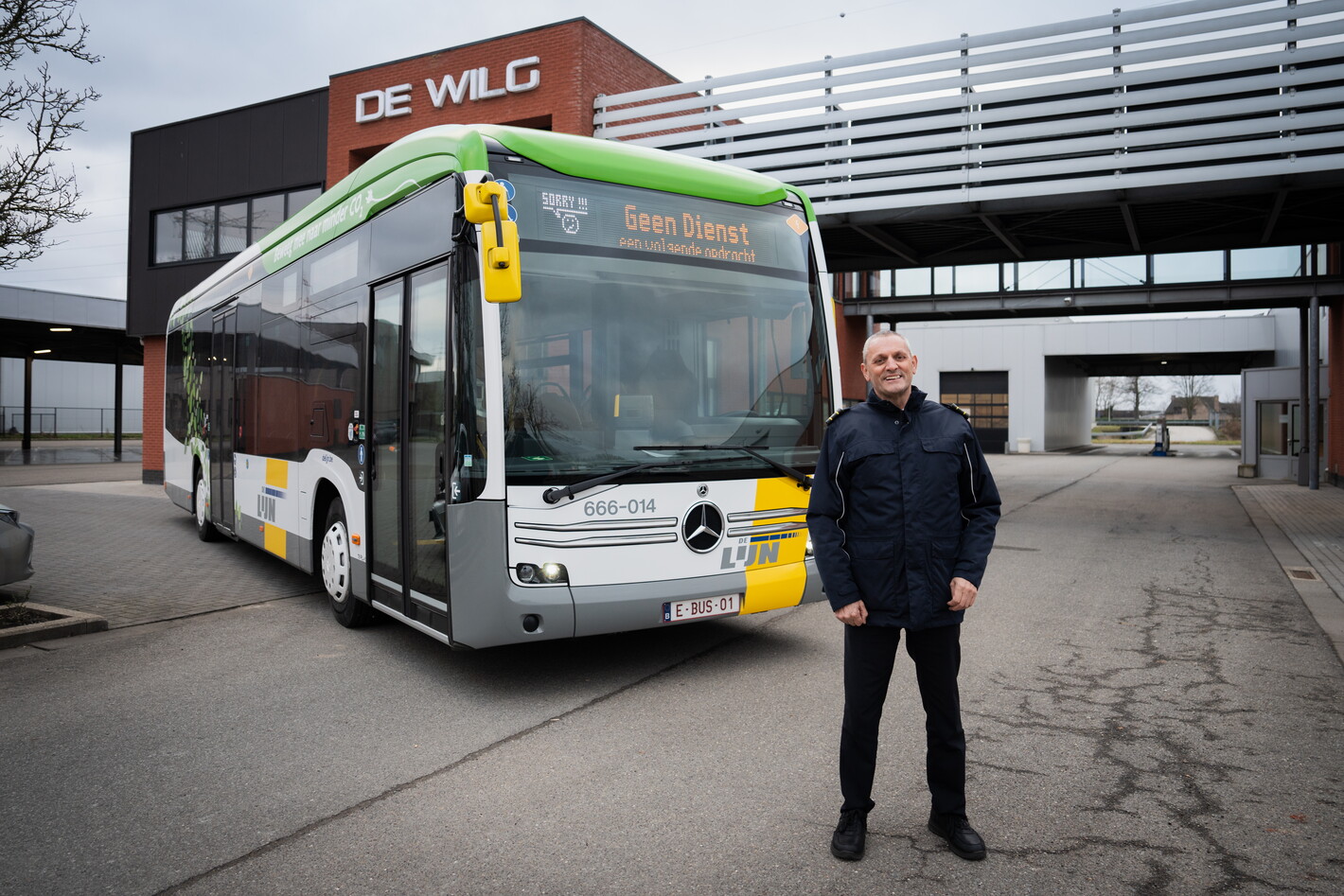 Man staat voor stadsbus bij station, bewolkte dag.