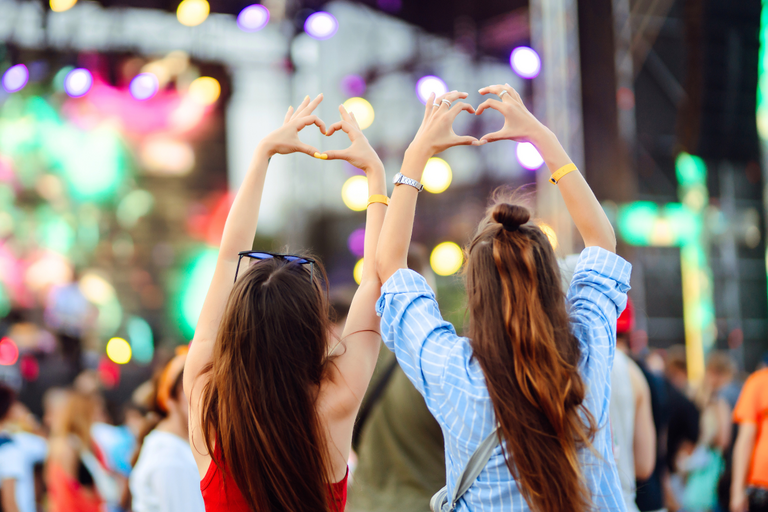 Twee vrouwen maken hartje met handen op een druk, kleurrijk festival.