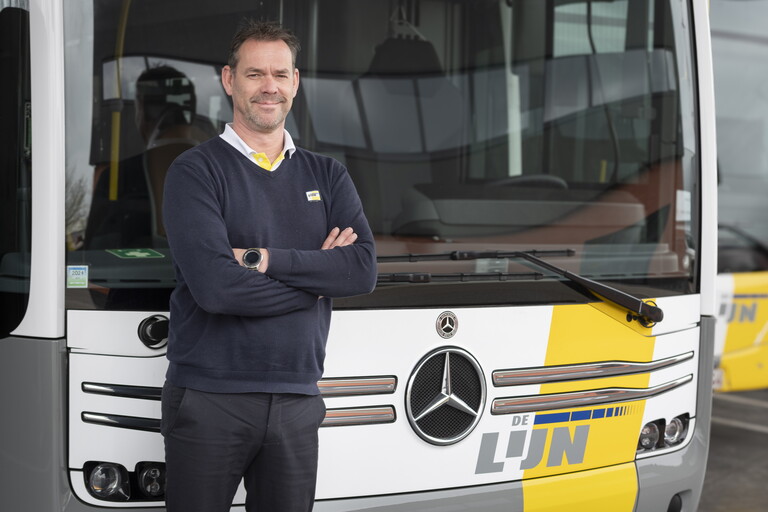 Man in uniform staat met gekruiste armen voor een gele vrachtwagen met Mercedes-logo.